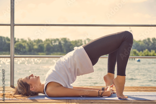 Woman in white undershirt and black leggings is practicing yoga performing yoga-asanas on a mat outdoors, near a riverside.