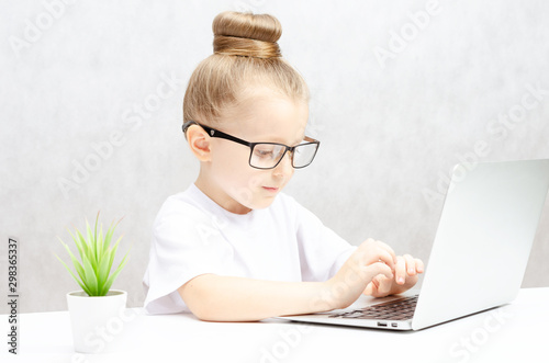 Little blonde girl in black glasses on a white background, at a white table, works on a laptop.