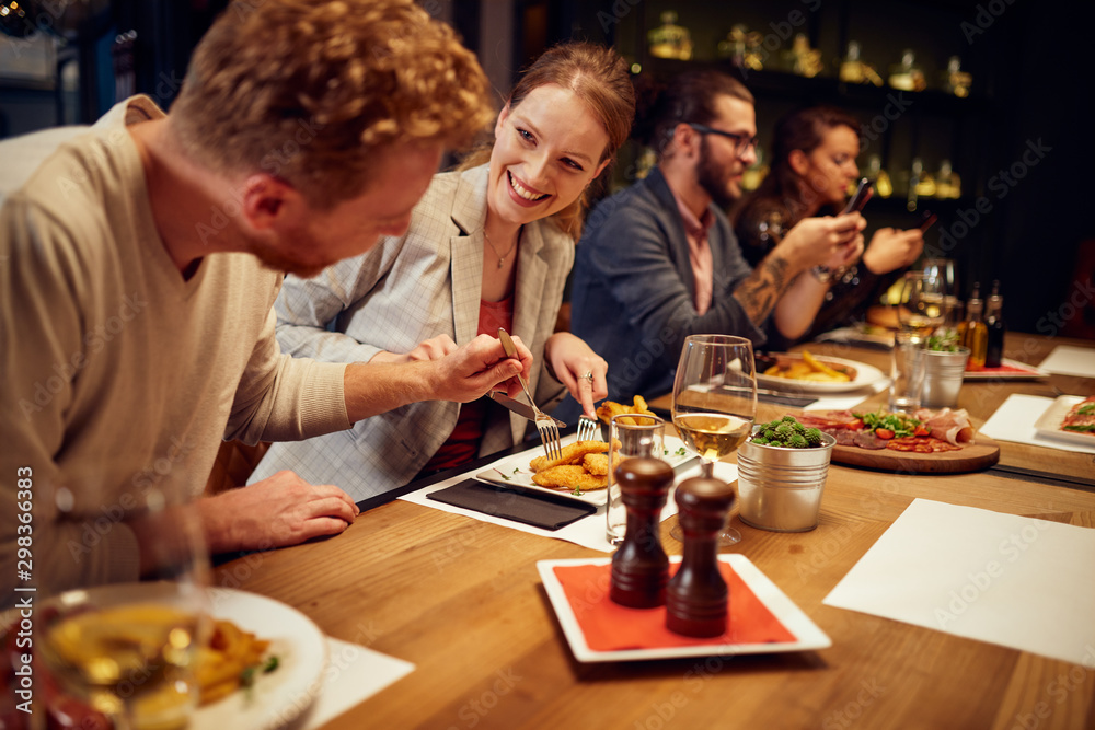 Handsome caucasian ginger taking food out of his girlfriend's plate while sitting in restaurant for dinner. In background are their friends. Handsome caucasian ginger taking food out of his girlfriend's plate while sitting in restaurant for dinner. In background are their friends.