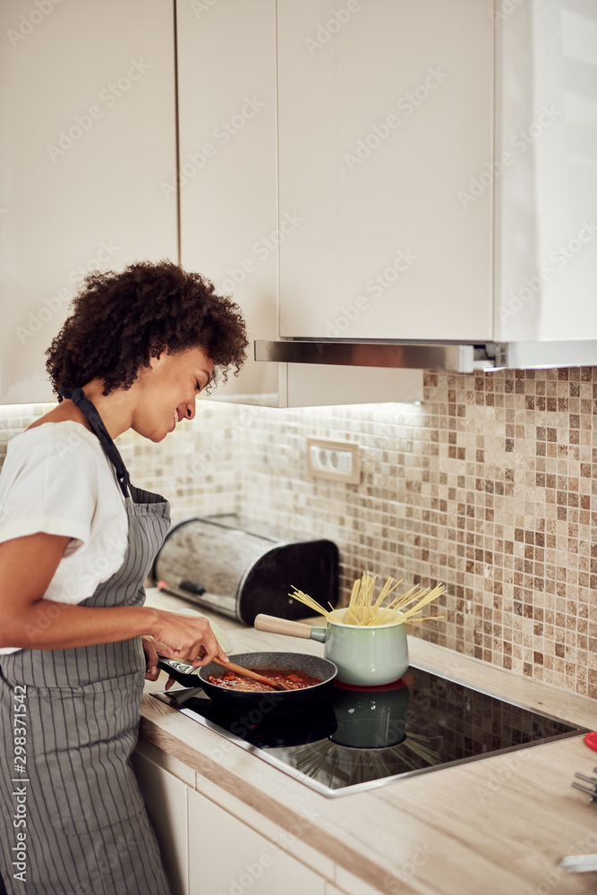 Mixed race woman in apron standing next to stove and stirring tomato sauce. On stove are saucepan and pot with spaghetti. Mixed race woman in apron standing next to stove and stirring tomato sauce. On stove are saucepan and pot with spaghetti.