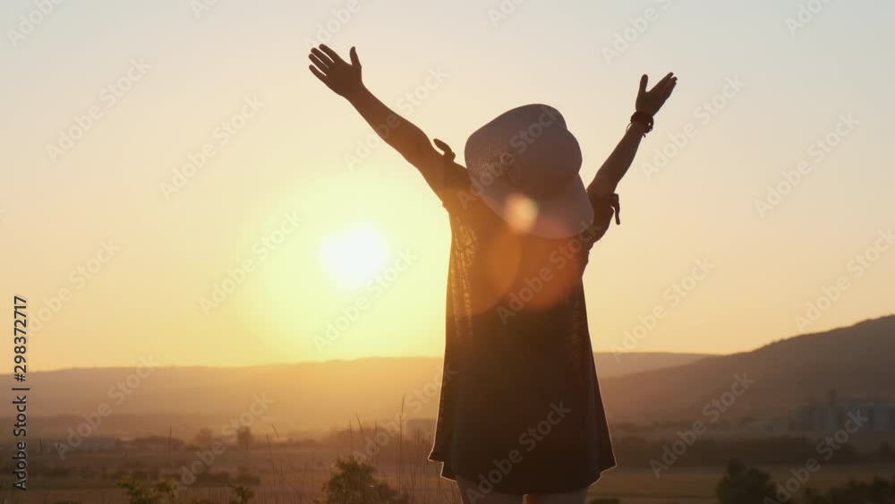 Slow motion silhouette happy woman in dress and hat standing in beautiful landscape field on background of bright sunset and admiring nature, raising her hands to the sun. Lens flare. Slow motion silhouette happy woman in dress and hat standing in beautiful landscape field on background of bright sunset and admiring nature, raising her hands to the sun. Lens flare.