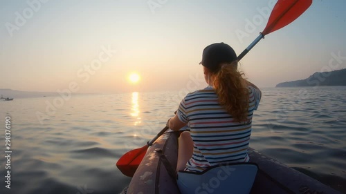 Happy young woman with her dog Jack Russell Terrier paddling on an inflatable kayak water lake a large mountain lake against orange sunrise slow motion slide. Family Sports Weekend. Lens flare. Gopro