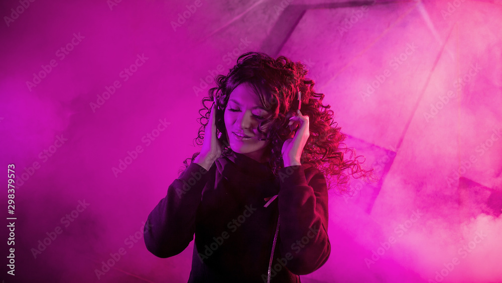 Young beautiful curly woman in big headphones smiles, listens to music, sings along, dances, jumps, closing her eyes.