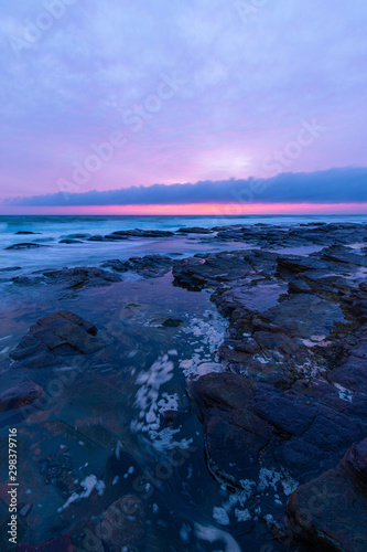Water flowing between rocks in the coastline with cloudy sunrise sky.
