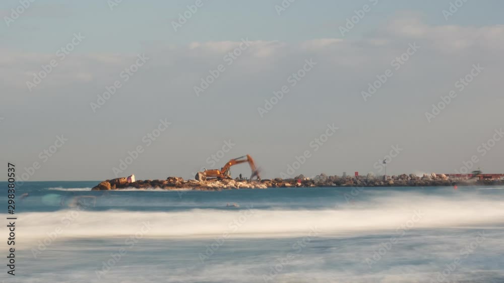 Timelapse of construction work being done early in the morning, on a breakwater. Blured waves in foreground and partly cloudy, mediterranean sky in the background. Timelapse of construction work being done early in the morning, on a breakwater. Blured waves in foreground and partly cloudy, mediterranean sky in the background.