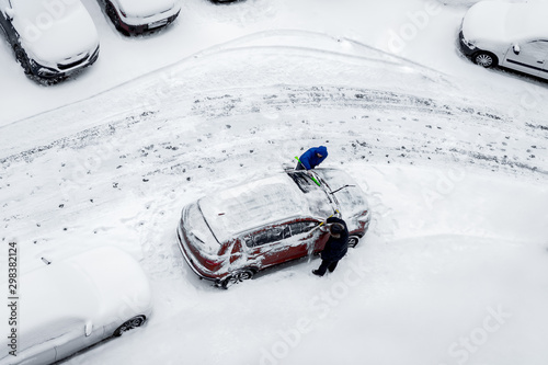 Two men, with brushes, remove snow from the car. The rest of the cars are covered with an even layer of snow. Top view, aerial photography. Winter daytime.