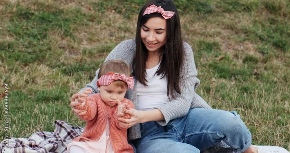 Mother and a small daughter, spends time together in a city park on a picnic. Young woman and little girl are playing on a plaid in green grass, at sunset Mother and a small daughter, spends time together in a city park on a picnic. Young woman and little girl are playing on a plaid in green grass, at sunset