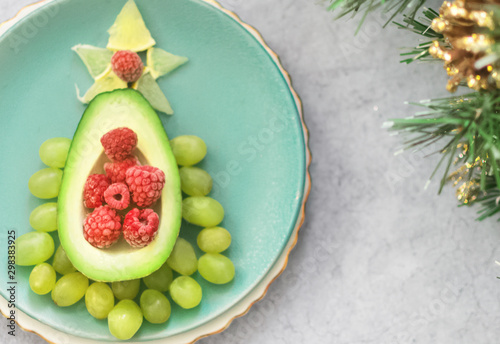 Delicious edible Christmas tree of avocado, frozen sweet raspberries and grapes on a blue plate on the table near the branches of the Christmas tree.Children's Breakfast.new year festive table
