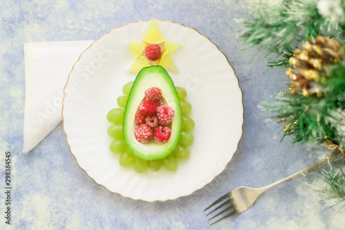 Delicious edible Christmas tree of avocado, frozen sweet raspberries and grapes on a white plate on the table near the branches of the Christmas tree.Children's Breakfast.new year festive table