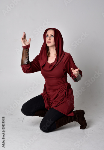 full length portrait of a brunette girl wearing a red fantasy tunic with hood. Sitting pose on a white studio background.