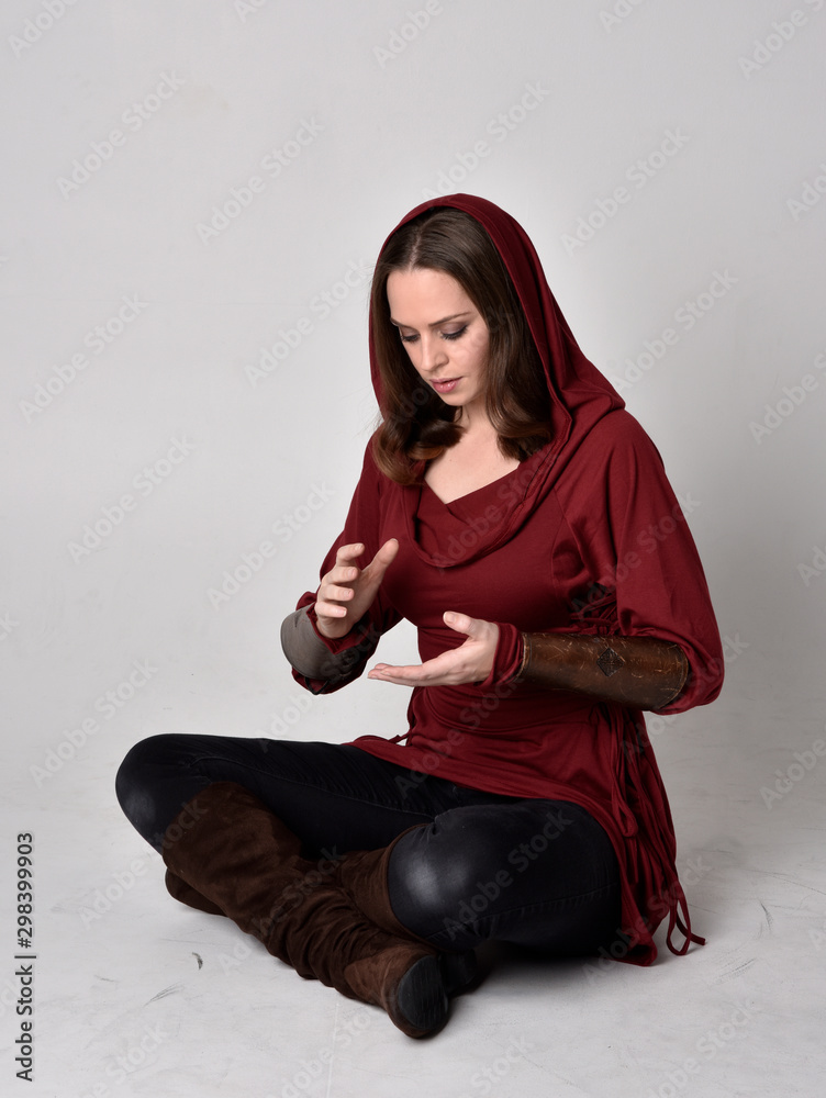 Fototapeta premium full length portrait of a brunette girl wearing a red fantasy tunic with hood. Sitting pose on a white studio background.