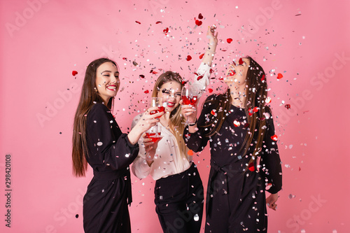Three women celebrate the New Year party having fun laughing under the flying confetti and drinking wine.