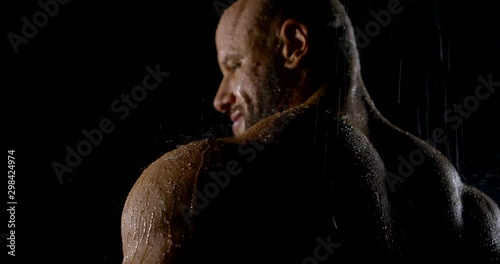 naked male culturist is standing under water flows in darkness, back view on big shoulders photo