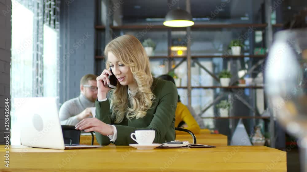 Tracking shot of smiling blonde businesswoman speaking on mobile phone and using laptop computer at cafe table on lunch