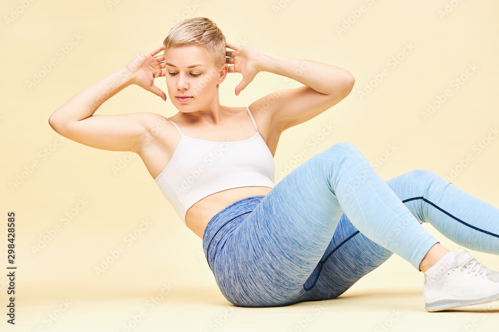 Gorgeous young European female fitness instructor with pixie blonde hair sitting on floor with hands behind her head, showing right technique of abdiminal crunches or crisscross exercise for toned abs Gorgeous young European female fitness instructor with pixie blonde hair sitting on floor with hands behind her head, showing right technique of abdiminal crunches or crisscross exercise for toned abs