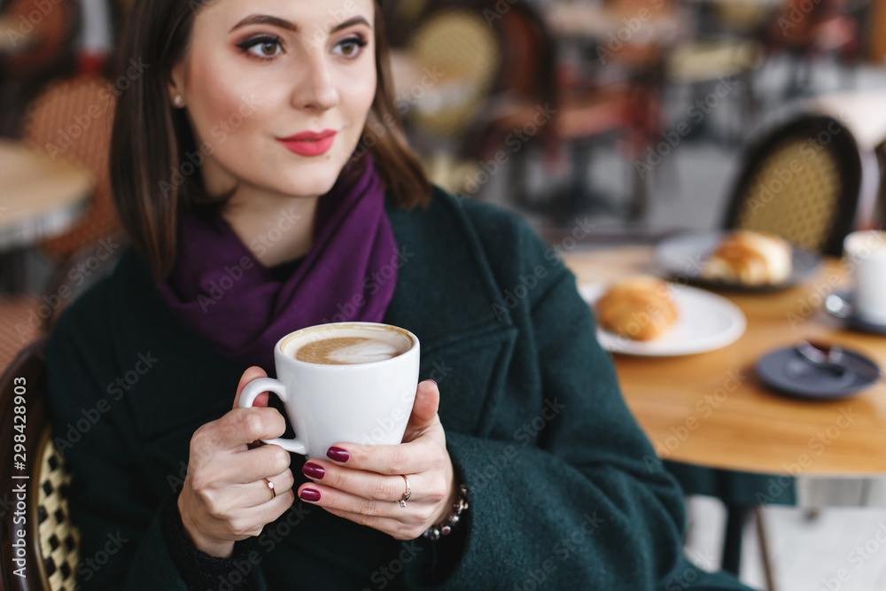 Beautiful young brunette woman with short hair wearing green coat and purple scarf sitting at a table in cozy street outdoor cafe and drinking coffee. Girl with cup of cappuccino at restaurant terrace Beautiful young brunette woman with short hair wearing green coat and purple scarf sitting at a table in cozy street outdoor cafe and drinking coffee. Girl with cup of cappuccino at restaurant terrace