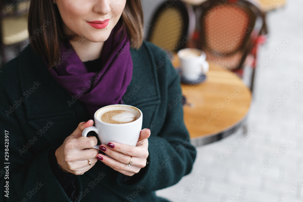 Cup of coffee in women's hands. Beautiful young brunette woman wearing green coat sitting at a table in cozy street outdoor cafe and drinking coffee. Girl with cup of cappuccino at restaurant terrace. Cup of coffee in women's hands. Beautiful young brunette woman wearing green coat sitting at a table in cozy street outdoor cafe and drinking coffee. Girl with cup of cappuccino at restaurant terrace.