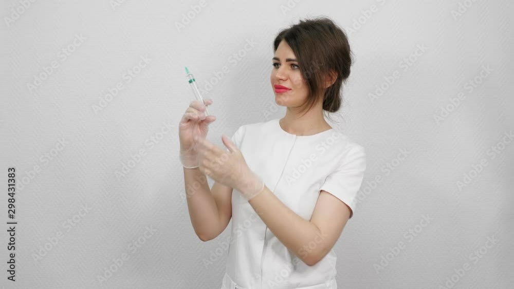 pretty woman doctor with dark hair in protection gloves pushes and shakes syringe standing against white wall close view pretty woman doctor with dark hair in protection gloves pushes and shakes syringe standing against white wall close view
