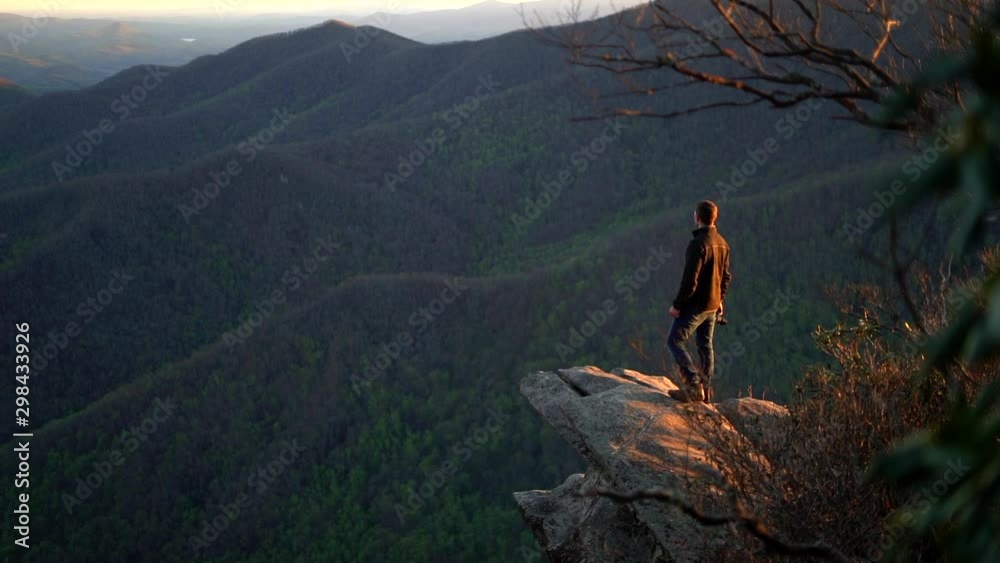 Hiking man is backlit by the setting sun as he stands on a cliff and looks out over the mountains and forest below