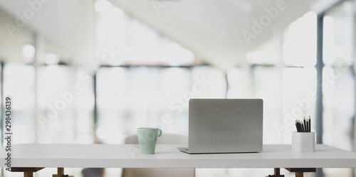 Laptop computer on white table and coffee cup in modern office with blurred background
