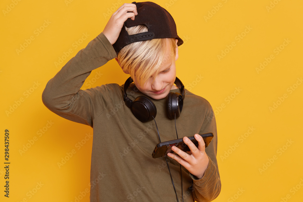 Little lond boy wearing in green sweater and blackcap, holding mobile phne  in hand, keeps hand on head, playing game on smart phone isolated over  yellow background. Kids childhood lifestyle concept. Photos
