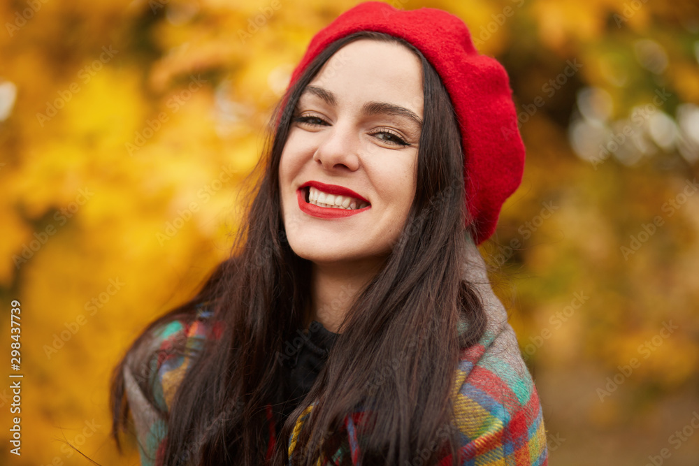 Autumn portrait of happy lovely and beautiful Caucasian young woman in forest in fall colors, attractive female dresses red beret and wrapped checkered blanket, poses smiling in autumn park.