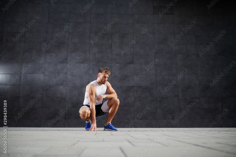 Young handsome sporty caucasian man in shorts and t-shirt crouching outdoors in front of gray wall. Young handsome sporty caucasian man in shorts and t-shirt crouching outdoors in front of gray wall.