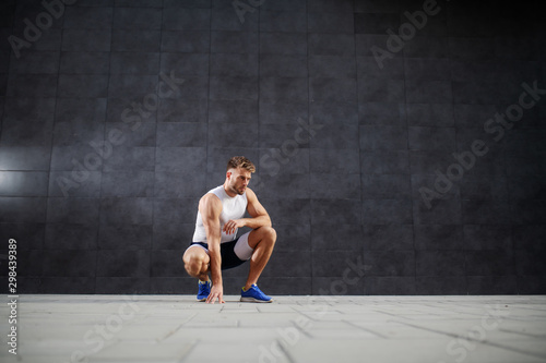 Young handsome sporty caucasian man in shorts and t-shirt crouching outdoors in front of gray wall.