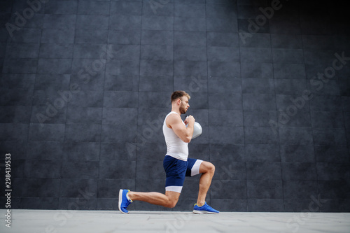 Handsome strong muscular caucasian man in shorts and t-shirt doing lunges and holding kettle bell. In background is gray wall.