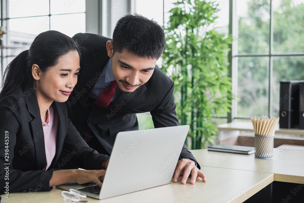 Confidence young Asian business man in suit leaning over a businesswoman typing on laptop working together at a bright office desk. Confidence young Asian business man in suit leaning over a businesswoman typing on laptop working together at a bright office desk.
