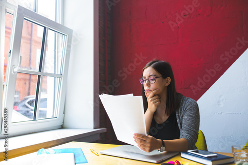 Serious successful business woman in stylish glasses reading paper documents while sitting at desktop in office interior. Female executive director reading resume during work day in company