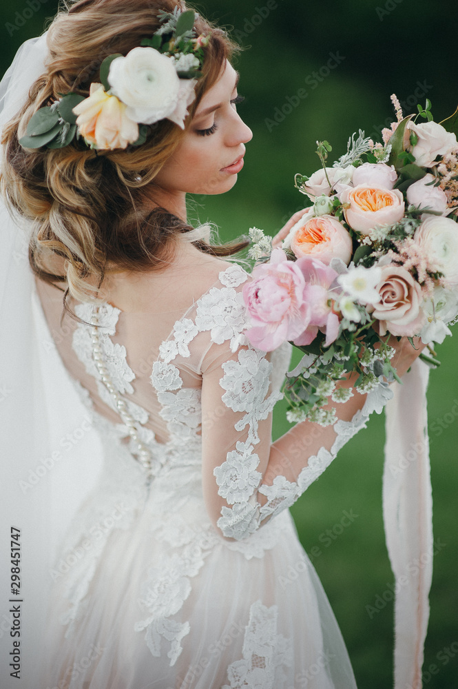 Naklejka premium Portrait of a beautiful bride with fresh flowers in her hair and a wedding bouquet of flowers. Posing on a background of green fields