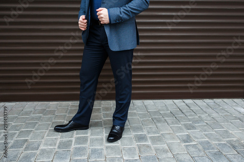 Man's legs. Man wearing suit jacket, pants, blue shirt and black shoes standing near brown roller door on the city street. Details of classic elegant formal look. Trendy men's outfit. Street fashion.