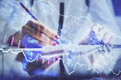 A woman hands writing information about stock market in notepad. Forex chart holograms in front. Concept of research.