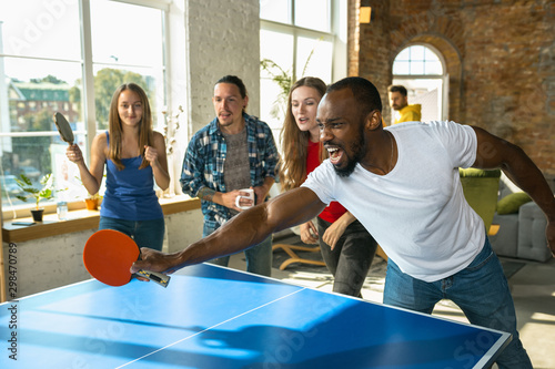 Young people playing table tennis in workplace, having fun. Friends in casual clothes play ping pong together at sunny day. Concept of leisure activity, sport, friendship, teambuilding, teamwork.