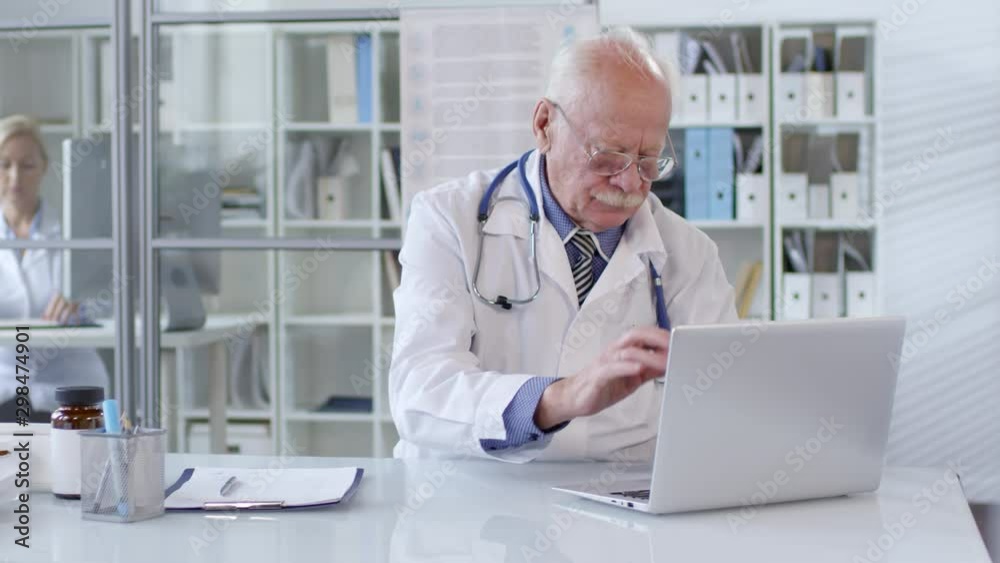 Portrait of senior doctor in eyeglasses and white coat working on laptop at desk and then looking at camera and smiling while female assistant working behind glass wall in the background