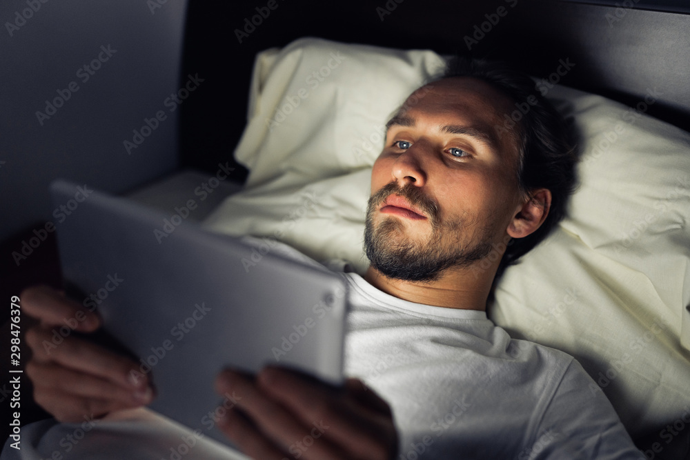 Young handsome and tired man with a beard cannot sleep and is watching something on his tablet at night.