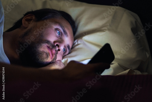 Young handsome and tired man with a beard cannot sleep and is watching something on his phone at night.