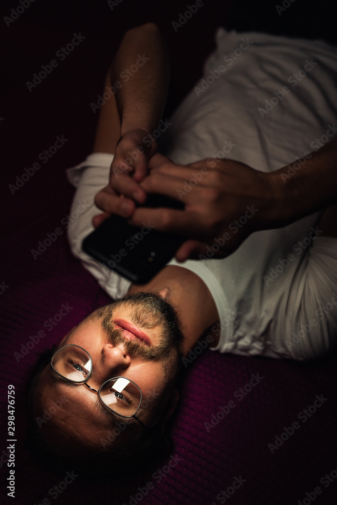 Bearded young man is lying in his bed at night while watching something on his mobile phone. Phone's reflection is visible in man's glasses.