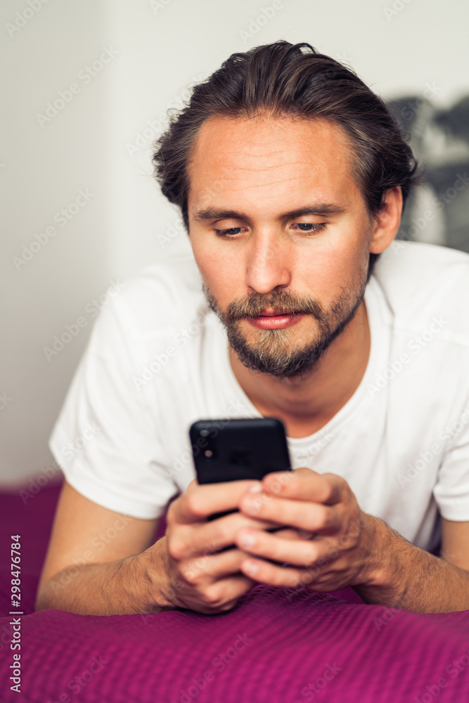 Rather serious young man that is lying in bed while looking at his mobile phone Rather serious young man that is lying in bed while looking at his mobile phone