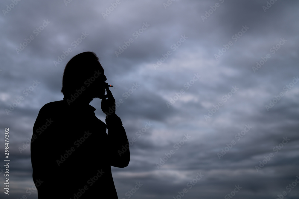 Dark silhouette of a handsome young man smoking a cigarette. Dark clouds visible in the background.
