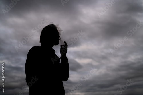 Dark silhouette of a handsome young man smoking a cigarette. Dark clouds visible in the background.