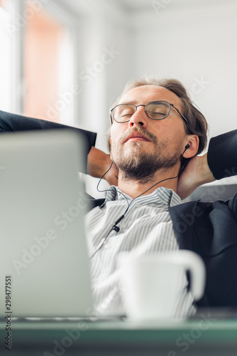 Young man is trying to relax and sleep while listening to music at work. Coffee cup and a computer visible in the foreground.