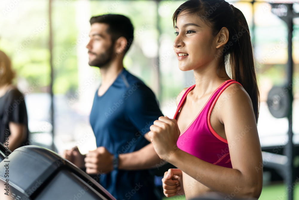 Fototapeta premium Young fit beautiful Asian woman and handsome Caucasian man running on treadmill or running machine in modern fitness gym. Seen from side view while they focusing on running. Workout in Gym and fitness