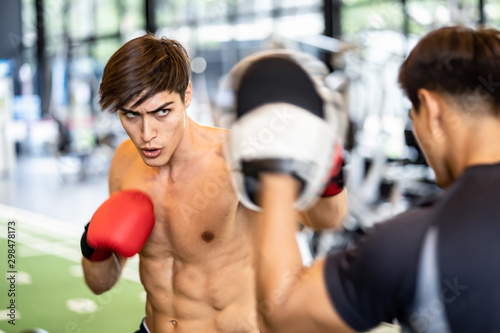 Soft focus shows moving action of Caucasian handsome man wearing boxing gloves punching ahead with trainer in gym or fitness club. Bear chested nice body boxing male looking forward with serious face.
