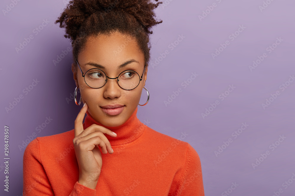 Photo of thoughtful African American woman keeps index finger on cheek, looks aside with pensive expression, thinks what to do, wears round eyewear, silver earrings and poloneck, models indoor - obrazy, fototapety, plakaty 