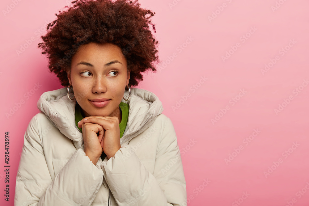 Attractive woman with serious look, dressed in outerwear, holds both hands together under chin, looks away, tries to warm during cold winter weather, models over pink background, copy space area Attractive woman with serious look, dressed in outerwear, holds both hands together under chin, looks away, tries to warm during cold winter weather, models over pink background, copy space area