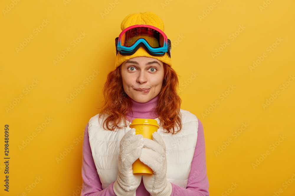 Lovely young woman bites lips, has foxy hair, bites lips, drinks hot beverage, wears ski goggles, looks directly at camera, poses against yellow background, has rest after training in winter forest Lovely young woman bites lips, has foxy hair, bites lips, drinks hot beverage, wears ski goggles, looks directly at camera, poses against yellow background, has rest after training in winter forest