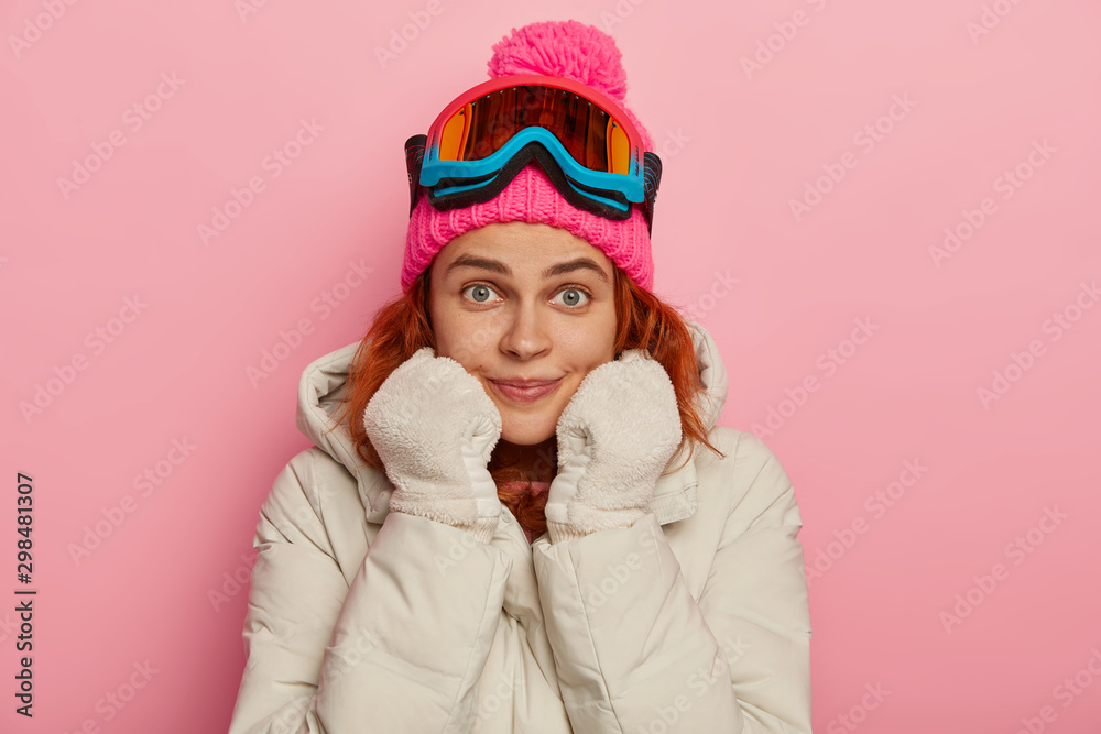 Beautiful ginger woman in winter clothing, enjoys outdoor sport, keeps both hands in mittens under chin, looks surprisingly at camera with green eyes, models against pink background. Pastime Beautiful ginger woman in winter clothing, enjoys outdoor sport, keeps both hands in mittens under chin, looks surprisingly at camera with green eyes, models against pink background. Pastime