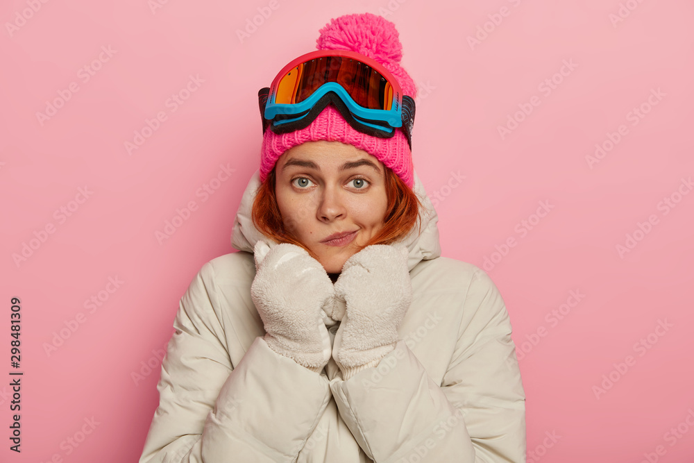 Photo of lovely European female traveler purses lips, warms herself in comfortable outerwear during winter time, wears ski mask, enjoys sunny weather in mountains and leasure outdoor activities